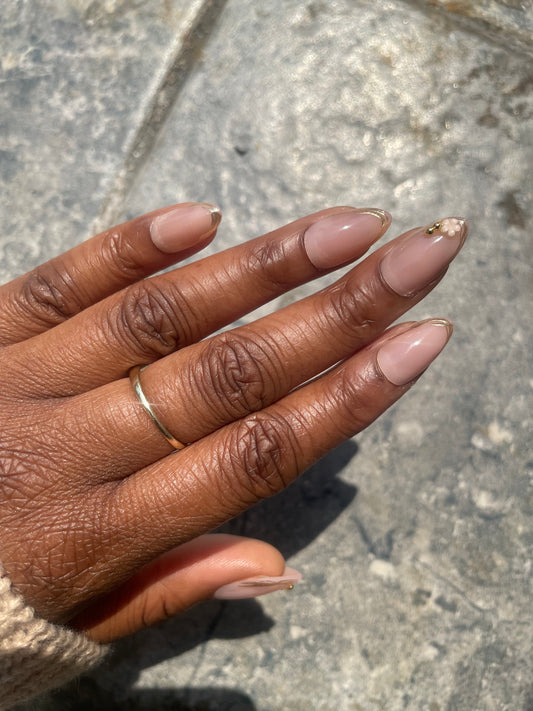 Close- up Camellia soft gel nude almond press-on with gold French tips and delicate floral accent on brown skin.